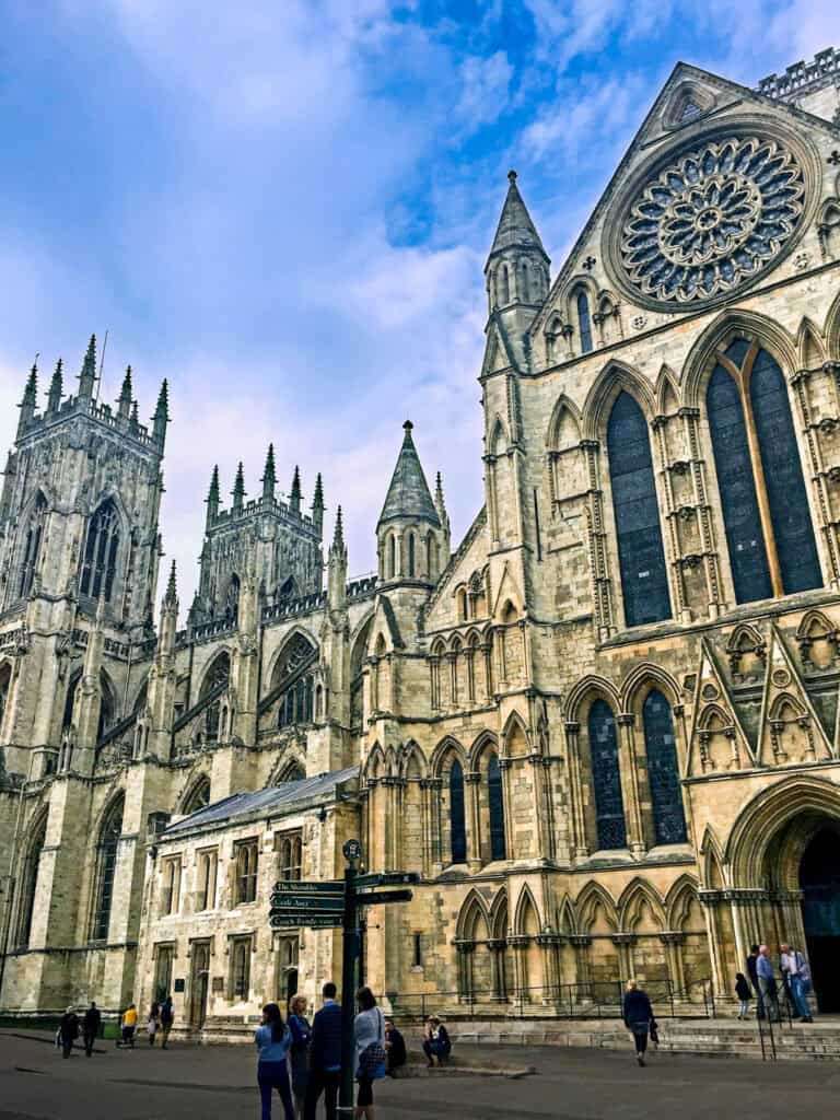 York Minster's Gothic cathedral with intricate stone facade, large stained glass window, and tall spires under a blue sky. People walk along the foreground.