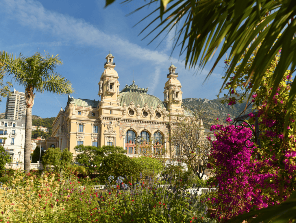 Monaco Casino building with towers and a green dome, surrounded by lush gardens with colorful flowers and palm trees under a clear blue sky.