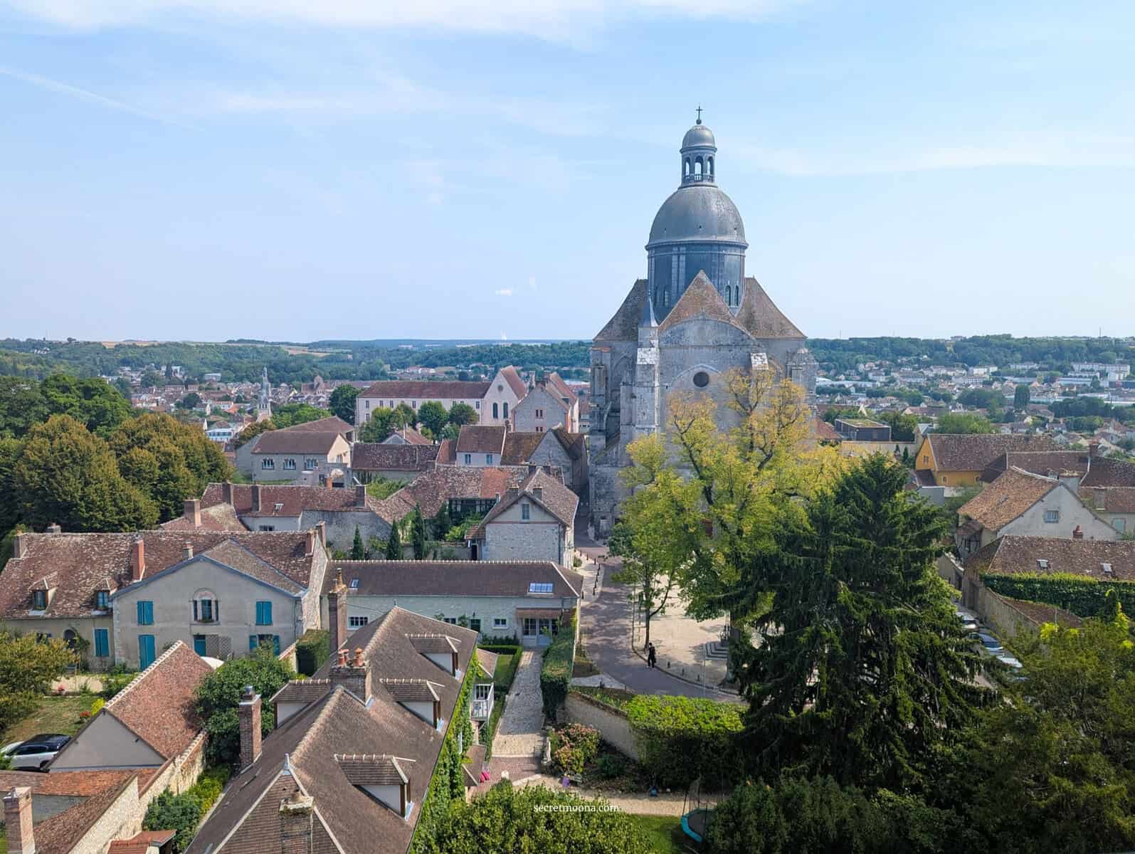 Aerial view of a historic Provins with a prominent church tower surrounded by charming houses and lush greenery.