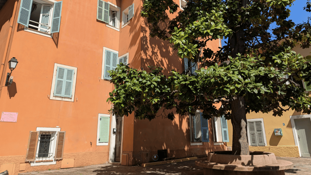 How To Spend One Day In Villefranche-sur-Mer: Things To Do 4 Quiet square in Villefranche-sur-Mer with a leafy tree, pastel buildings and outdoor seating