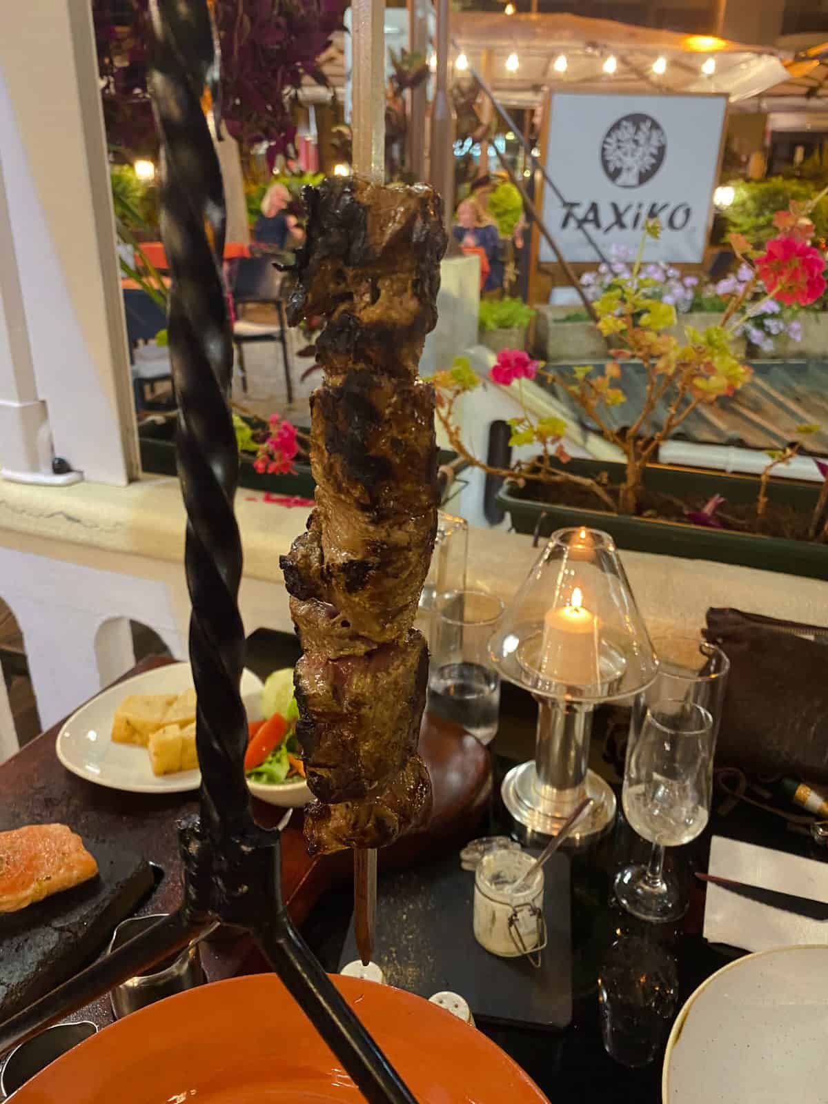 Beef espetada threaded onto a laurel skewer and hung from a metal stand on a restaurant table