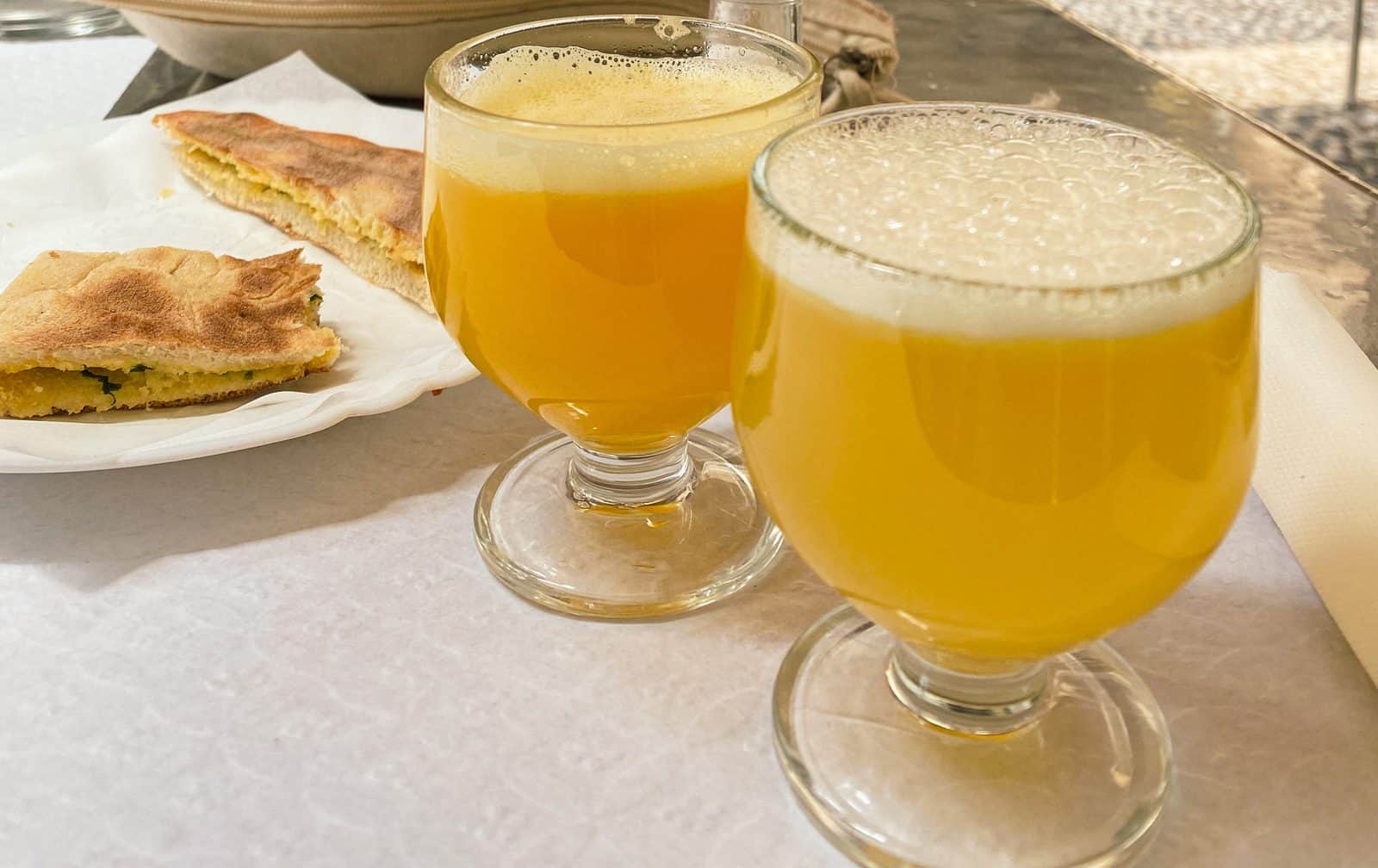 Two glasses of cloudy orange poncha beside a plate of bolo do caco flatbread