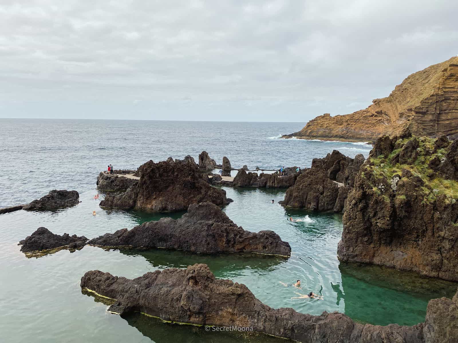 The volcanic rock pools at Porto Moniz filled with turquoise seawater, with waves breaking on black lava beyond