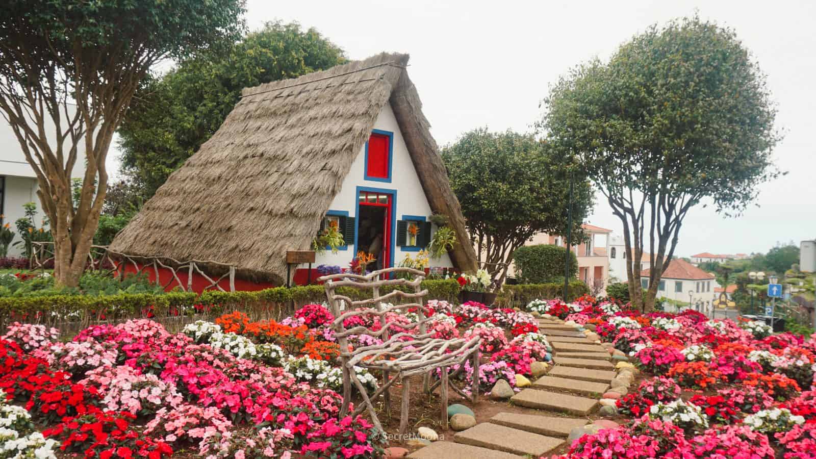 A traditional triangular thatched house in Santana, painted white with red and blue trim, surrounded by flowers