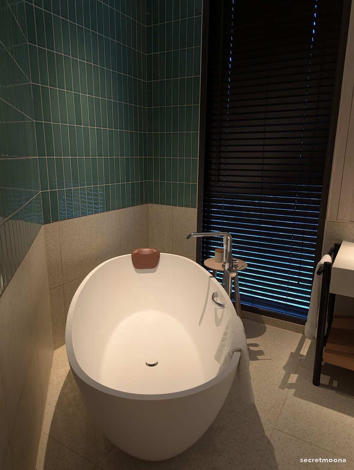 Freestanding egg-shaped bathtub against green tiling beside louvred shutters
