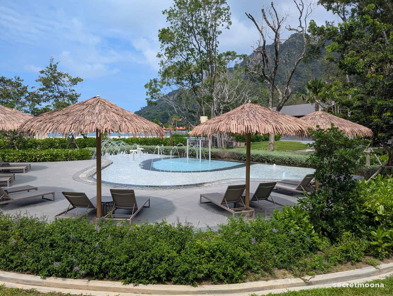 A family pool with thatched parasols and loungers, backed by jungle and mountains